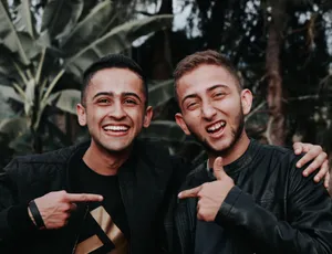 Two young men smiling and pointing at each other in an outdoor setting with lush greenery. jigsaw puzzle