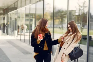 Young smiling best girlfriends in casual wear looking at each other and having fun while standing near glass building in city street jigsaw puzzle