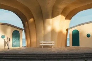 Woman stands under archway with buildings, bench, and ocean view. jigsaw puzzle
