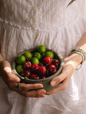 A woman in white dress holds a rustic bowl with red and green cherries. jigsaw puzzle