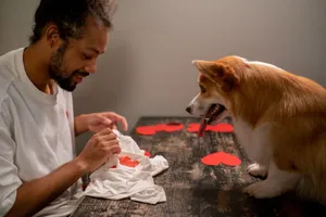A man crafts heart decorations on a table indoors with his attentive dog nearby. jigsaw puzzle