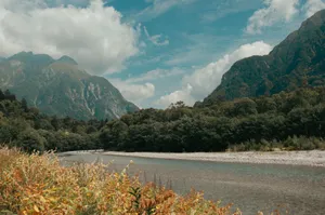 Serene river scene with lush green trees and towering mountains under a clear sky. jigsaw puzzle