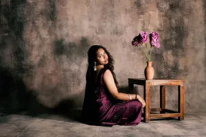 A woman in a purple dress sits gracefully beside a wooden table with a vase of flowers. jigsaw puzzle