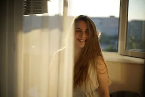 A young woman smiles warmly by a window as sunlight streams through a curtain. jigsaw puzzle