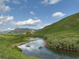 Tranquil river flowing through lush green hills under a blue sky with clouds. jigsaw puzzle