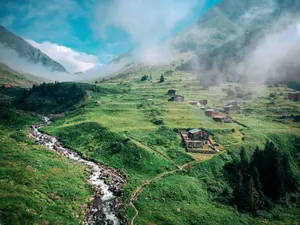 Idyllic mountain valley with green hills, flowing stream, and quaint houses under misty clouds. jigsaw puzzle