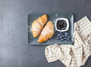 A top view of a breakfast arrangement featuring croissants, blueberries, and jam on a checkered cloth. jigsaw puzzle