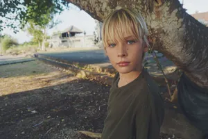 A blonde boy standing outdoors by a tree, capturing a quiet moment. jigsaw puzzle