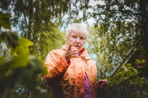 Senior woman outdoors pointing amidst greenery, wearing an orange jacket. jigsaw puzzle