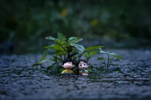 Two cute figurines sitting in the rain, surrounded by a small plant near a stream. jigsaw puzzle
