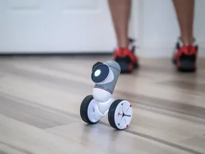 A close-up of a modern wheeled robot on a wooden floor with legs in the background. jigsaw puzzle