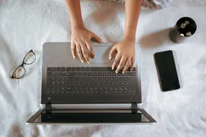 From above of crop unrecognizable female remote employee working on netbook near cellphone and eyeglasses on crumpled bed sheet jigsaw puzzle