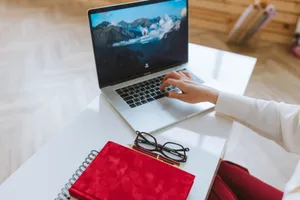 A person working on a laptop with a red notebook and glasses on a white table. jigsaw puzzle