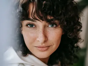 Headshot of attractive female with dark curly hair looking at camera against green leaves on blurred background in light room jigsaw puzzle